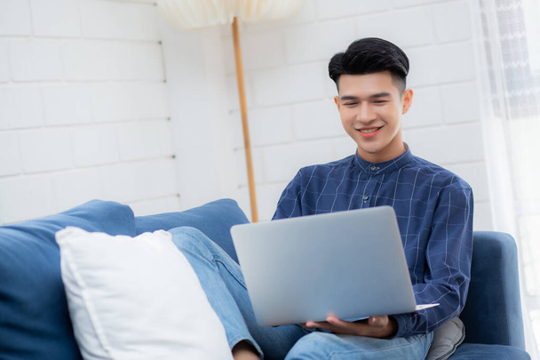 Young asian businessman smile and work from home with laptop computer online to internet on sofa in living room, freelance man using notebook on couch with comfort, new normal, lifestyle concept. - Photo, Image