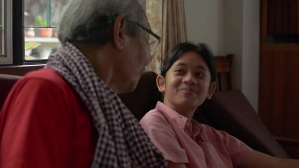 Schattige Aziatische tiener kleindochter lacht gelukkig terwijl ze zit en praat met haar senior grijze haar grootvader in de woonkamer. Verbinding van twee generaties in een familie. Relatie tussen ouderen en kind. - Video