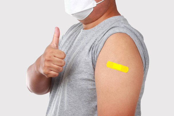 Beyaz maskeli Asyalı şişman bir adam gri bir gömlek giyiyor, sol kolunda sarı alçı var, COVID-19 (Coronavirus) aşısı gri bir arka planda duruyor. İzole. Kesme Yolu - Fotoğraf, Görsel