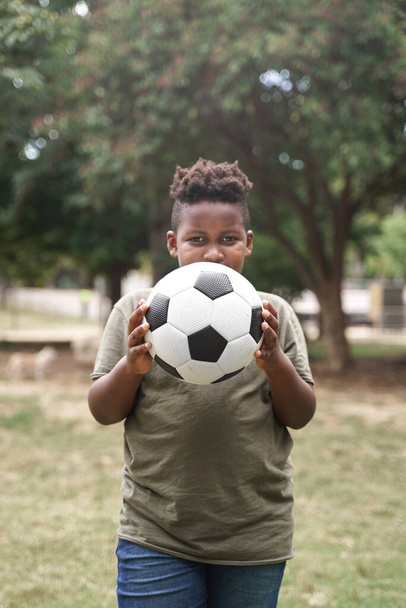 Αφρο-Αμερικανός που κρατάει μπάλα ποδοσφαίρου και καλύπτει το πρόσωπό του έξω στο πάρκο - Φωτογραφία, εικόνα