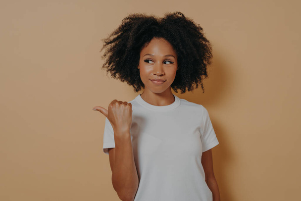 Hübsche schwarzhaarige Afrikanerin zeigt Daumen zur Seite, empfiehlt Werbung und wirbt für Produkt - Foto, Bild