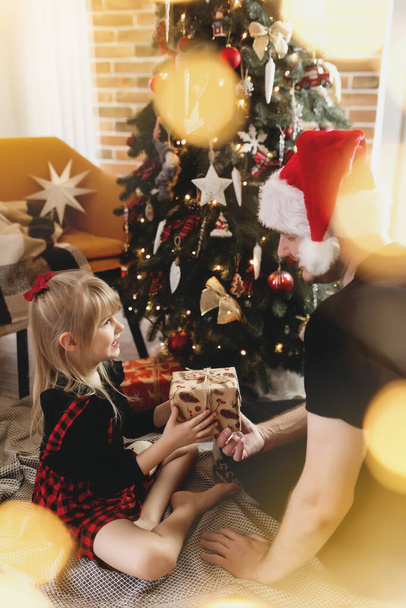 Kırmızı Noel Baba şapkalı genç baba süslü Noel ağacının yanındaki gülümseyen kızına Noel hediyesi veriyor. Kırmızı-siyah Noel kıyafeti giymiş bir kız. Gülümsüyorlar, yeni yıldan dolayı mutlular. - Fotoğraf, Görsel