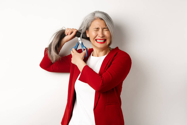 Bang aziatische vrouw knippen haar met een schaar en zich angstig voelen, sluit de ogen zenuwachtig, staan in rode blazer tegen witte achtergrond - Foto, afbeelding