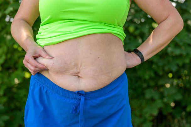 Donna in sovrappeso pizzicando il grasso in eccesso sullo stomaco tra le dita in un primo piano vista del suo midriff in abbigliamento sportivo - Foto, immagini
