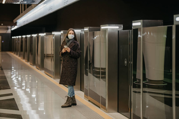 Μια γυναίκα με μάσκα προσώπου για να αποφύγει την εξάπλωση του κορωναϊού περιμένει το τρένο και κρατάει ένα smartphone στο σταθμό του μετρό. Ένα κορίτσι με χειρουργική μάσκα κρατάει κοινωνική απόσταση στο μετρό.. - Φωτογραφία, εικόνα
