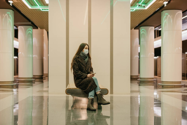 コロナウイルスの拡散を避けるために医療用フェイスマスクの孤独な女性が地下鉄のプラットフォーム上のスマートフォンで座っています。外科用マスクの少女が地下鉄で社会的距離を保っています. - 写真・画像