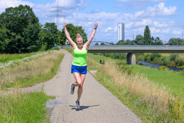 Happy vivacious sporty woman celebrating the hot summer weather while out enjoying a jog along a footpath beside a river cheering with arms raised in a healthy active lifestyle and vitality concept - Photo, Image