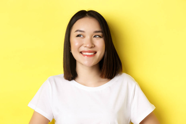 Közeli kép a jóképű ázsiai nő fehér pólóban, mosolygós és pillantás balra elégedett arc, áll a sárga háttér felett - Fotó, kép
