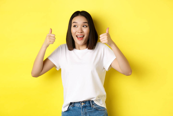 Красивая молодая женщина в белой футболке, показывая вверх большие пальцы и улыбаясь, хвалит хорошее предложение, рекомендует продукт, стоя довольная на желтом фоне - Фото, изображение