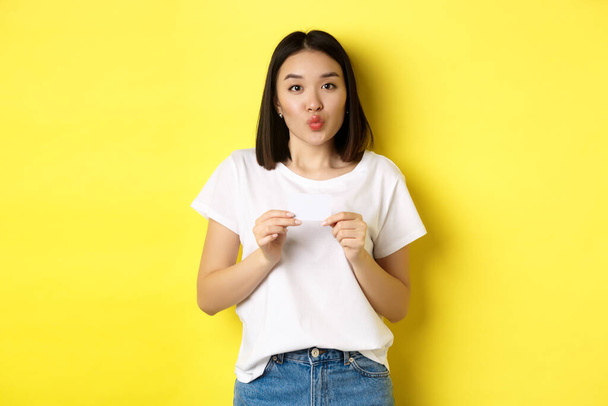 Young asian woman in casual white t-shirt showing plastic credit card and pucker lips silly, going on shopping, yellow background - 写真・画像