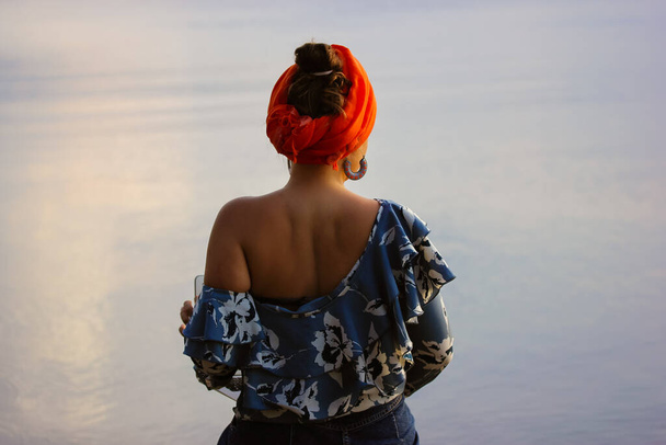Back view of a woman in a one-shoulder blouse with an orange turban sitting by the sea on a summer evening, twilight. Glaze on a blue water. Rest by the ocean, tranquility, harmony, calmness concept. - Foto, Bild