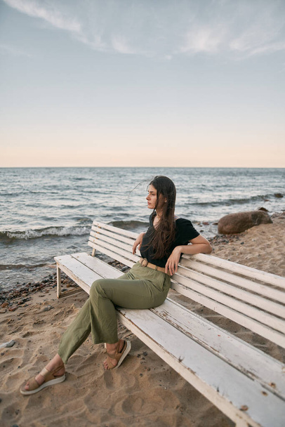 Spokój dziewczyna z ciemnymi włosami siedzi na białej ławce na brzegu morza. - Zdjęcie, obraz