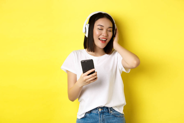 Cool asian girl singing along and listening music in wireless headphones, holding smartphone in hand, standing over yellow background - Photo, Image