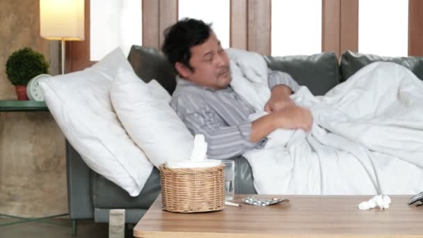 Hombre de oficina asiática está ausente del trabajo y licencia por enfermedad, toma la temperatura por un termómetro, toma medicamentos, y duerme en mantas en el sofá en casa, tejidos, y pastillas en la mesa. - Metraje, vídeo