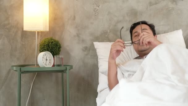 Hombre de oficina asiática está ausente del trabajo, se despierta tarde y baja por enfermedad, toma medicamentos, y duerme en mantas blancas en el sofá en casa, pañuelos y pastillas en la mesa frente a una pared de cemento gris. - Metraje, vídeo