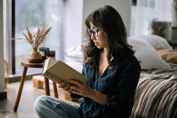 Young focused asian woman wearing eyeglasses reading book while sitting in lotus pose on floor in cozy living room decorated with dried wheat and candles, spending leisure time at home - Photo, Image