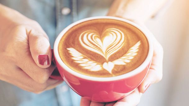 Κοντινό πλάνο ενός κόκκινου φλυτζανιού καφέ latte art σε γυναικείο χέρι σε καφετέρια - Φωτογραφία, εικόνα