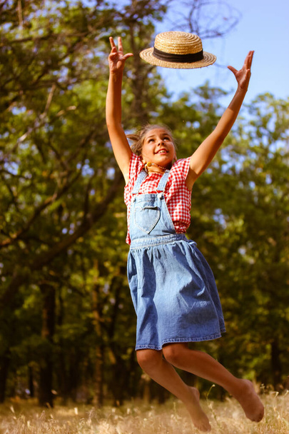 Una adolescente deportiva en un vestido de mezclilla con las manos levantadas salta por un sombrero de paja en un bosque de verano. Feliz infancia sin preocupaciones, recreación, concepto de alegría. Un niño de 10-12 años, niño juega al aire libre.  - Foto, imagen