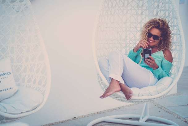 Pěkná dospělá žena sedět na bílém pohodlném sedadle ve venkovní zahradě s úsměvem a pomocí mobilního telefonu na video hovor  - Fotografie, Obrázek