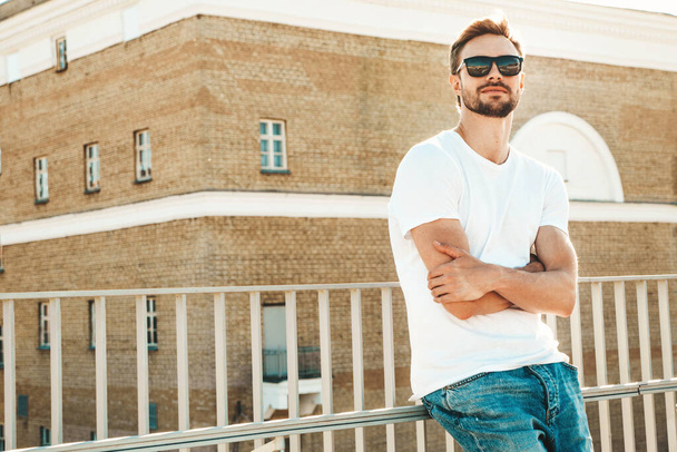 Portrait of handsome smiling  hipster lambersexual model.Stylish man dressed in white T-shirt and jeans. Fashion male posing on the street background near fence at the beach  - 写真・画像