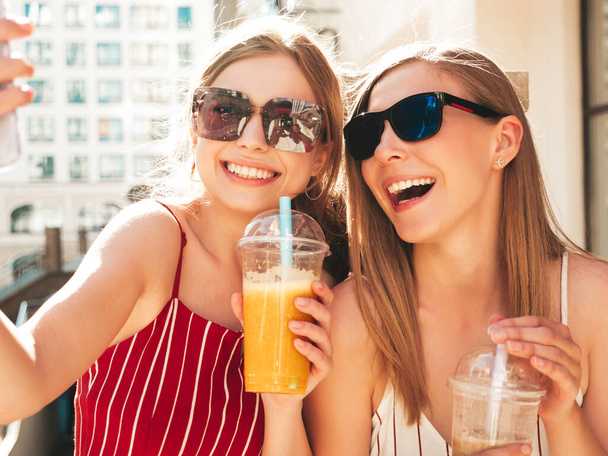 Two young beautiful smiling hipster female in trendy summer clothes. Carefree women posing outdoors.Positive models drinking fresh cocktail smoothie drink in plastic cup with straw.Taking selfie - Valokuva, kuva