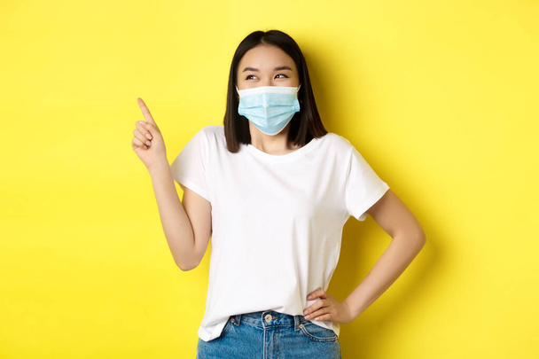 Covid, gezondheidszorg en pandemie concept. Aziatische vrouw in medisch masker en wit t-shirt wijzende vinger op de linker bovenhoek logo, met promotie, gele achtergrond - Foto, afbeelding
