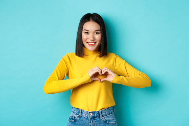 Valentinstag und Romantikkonzept. Schöne asiatische Frau zeigen, dass ich dich liebe, Herzgeste und Lächeln, vor blauem Hintergrund stehend - Foto, Bild