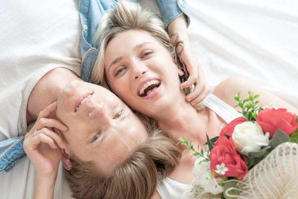ベッドの上に寝そべって花の花束とハンサムな男を保持している若いカップルの美しい女性のトップビューの肖像画,彼らは陽気でロマンチックな瞬間と笑みを浮かべています-愛のカップルのコンセプト - 写真・画像