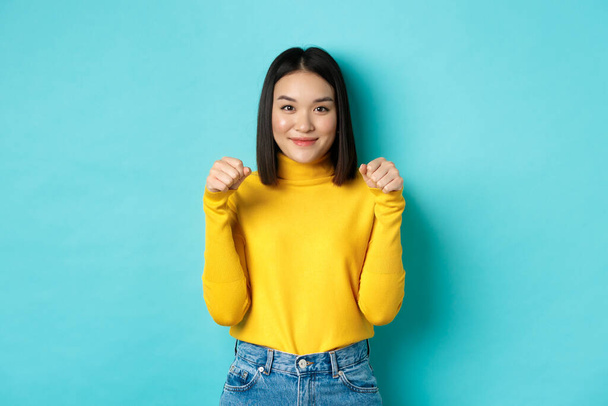 Piękno i moda. Piękna i stylowa azjatycka kobieta w żółtym pulowerze, trzymająca się za ręce w pobliżu klatki piersiowej, jakby trzymała sztandar lub logo, stojąca nad niebieskim tłem - Zdjęcie, obraz