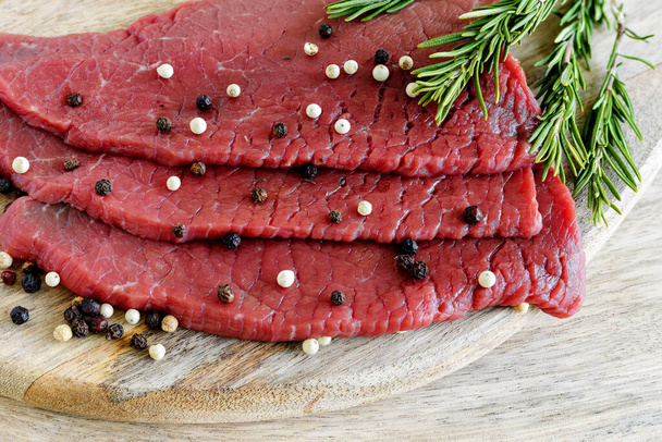 Raw slices of lean beef with pepper and rosemary on cutting board - Photo, image