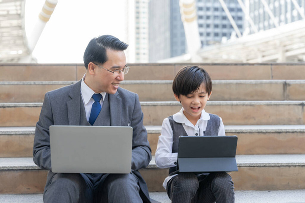 Отец-одиночка отец и его сын, используя ноутбук компьютер и планшетный телефон смартфон вместе на деловом районе города, папа и сын носить костюм бизнес семейной концепции - Фото, изображение