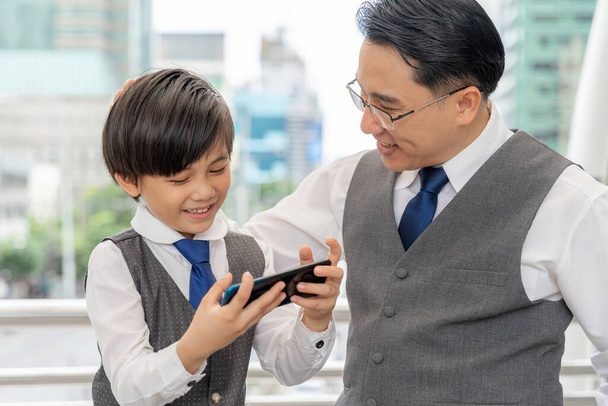 Πατέρας και γιος παίζουν έξυπνο τηλέφωνο μαζί στην επιχειρηματική περιοχή της πόλης, ο μπαμπάς και ο γιος ευτυχισμένη ασιατική οικογένεια έννοια - Φωτογραφία, εικόνα