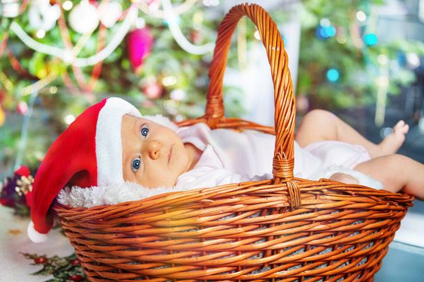 Beautiful baby girl in in the basket in Santa hat near Christmas tree - Fotó, kép