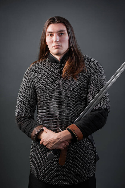 Portrait of a medieval warrior of the late Viking era and the beginning of the Crusades. Knight with long hair in chain mail, armed with a sword, isolated on dark background. - Photo, image