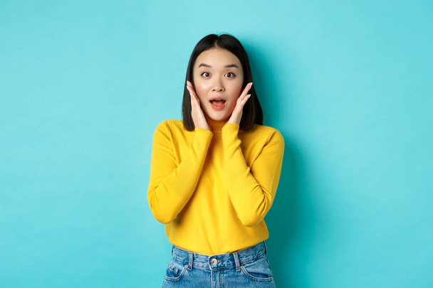 Retrato de chica asiática sorprendida mirando promo, jadeando asombrado y tocando mejillas, mirando fijamente a la cámara asombrado, fondo azul - Foto, imagen