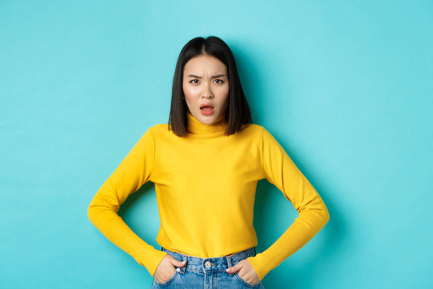 Portrait of disappointed korean woman in yellow sweater, frowning and looking upset, standing over blue background - 写真・画像
