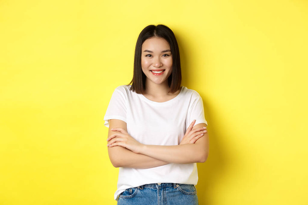 Magabiztos és elegáns ázsiai nő keresztbe karok mellkas és mosolygós, álló sárga háttér - Fotó, kép