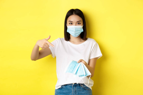Coronavirus, Quarantäne und Medizinkonzept. Junge asiatische Frau zeigt mit dem Finger auf medizinische Masken, um ins Freie zu gehen, steht über gelbem Hintergrund - Foto, Bild