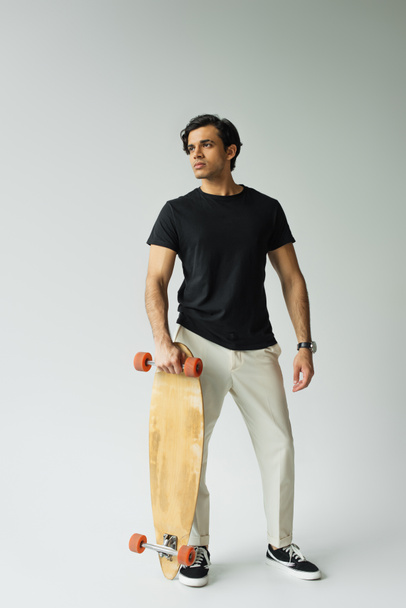 full length of young man in black t-shirt posing with longboard on grey - Zdjęcie, obraz