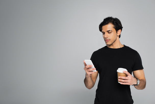 young man holding coffee to go and using smartphone isolated on grey - Photo, Image