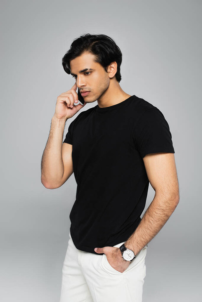 young man talking on cellphone and posing with hand in pocket isolated on grey - Photo, image
