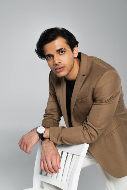 young man in beige blazer leaning on chair on grey - Foto, imagen