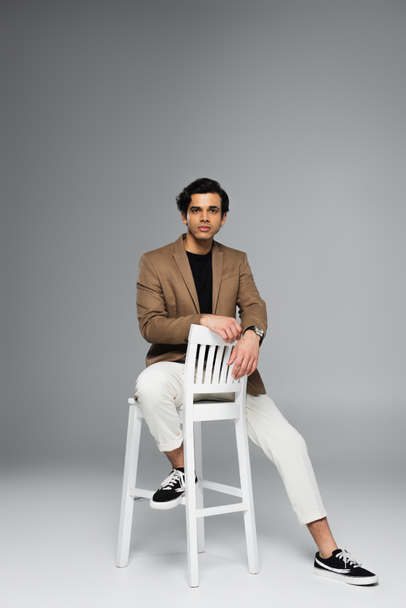 full length of young man in blazer sitting on white chair on grey - Zdjęcie, obraz