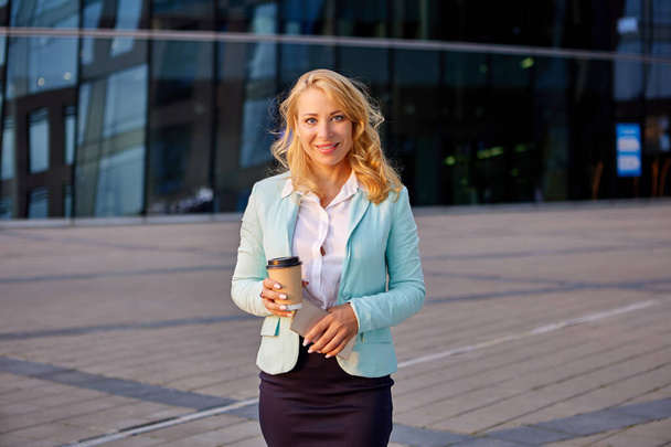 Cheerful businesswoman stands outdoors in business suit and with cup of coffee. - Fotografie, Obrázek