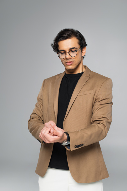 young man in stylish eyeglasses adjusting wristwatch isolated on grey  - Foto, afbeelding