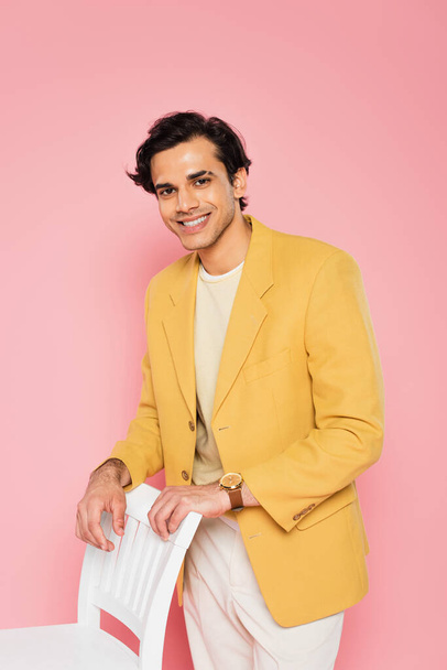 happy young man in yellow blazer leaning on white chair isolated on pink - Photo, Image