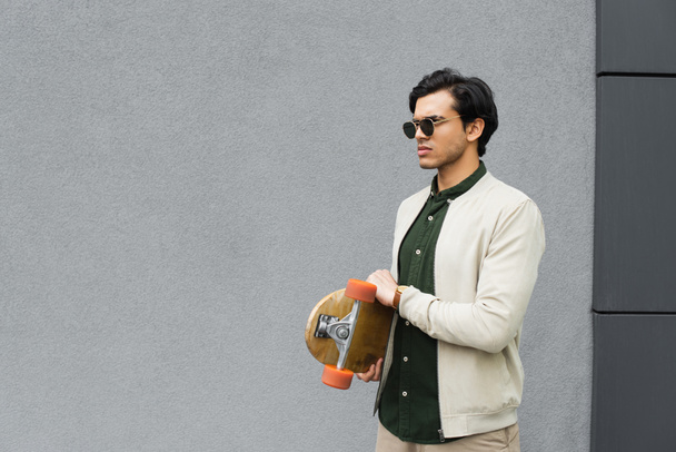 trendy young man in sunglasses holding longboard near building  - Фото, изображение