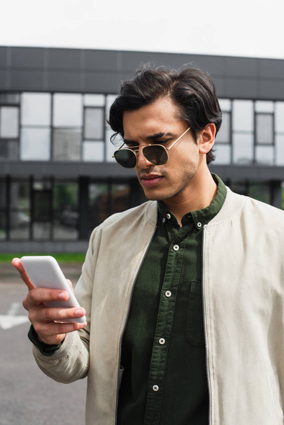 élégant jeune homme en lunettes de soleil et veste bombardier textos sur smartphone près du bâtiment  - Photo, image