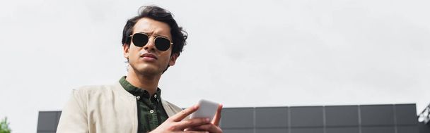 stylowy młodzieniec w okularach przeciwsłonecznych i bombowej kurtce trzymający smartfona na zewnątrz, baner - Zdjęcie, obraz