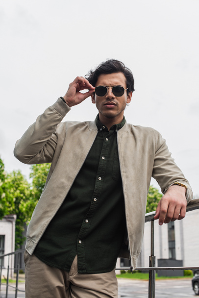 young man in bomber jacket adjusting stylish sunglasses while posing outside - Foto, imagen
